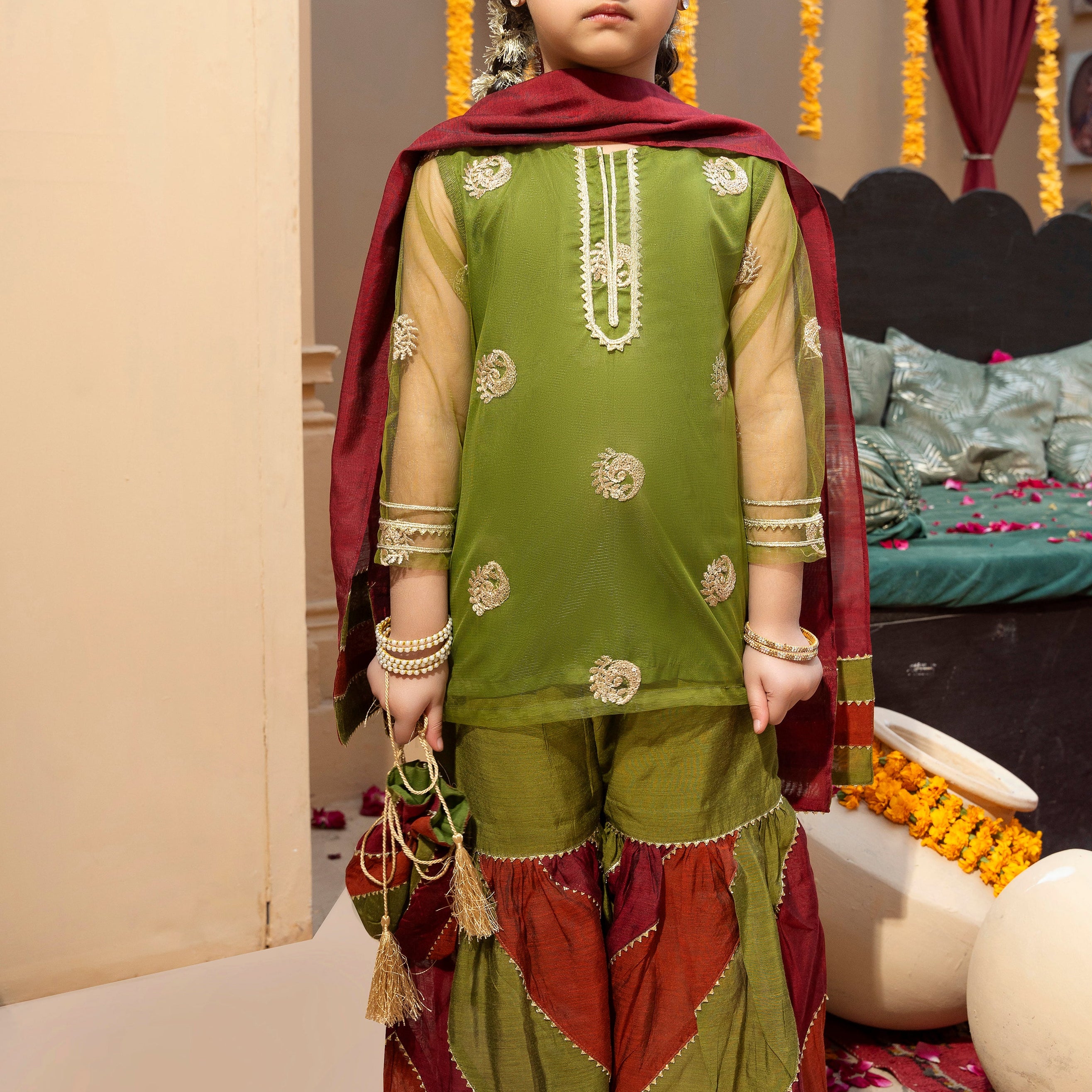 A young girl wearing a traditional 4-piece outfit with gold zari and sequins embroidery on a net shirt, chattapatti technique gharara in green, rust, and maroon with gold accents, and a matching dupatta with gold lace at the edges. She is also holding a potli.