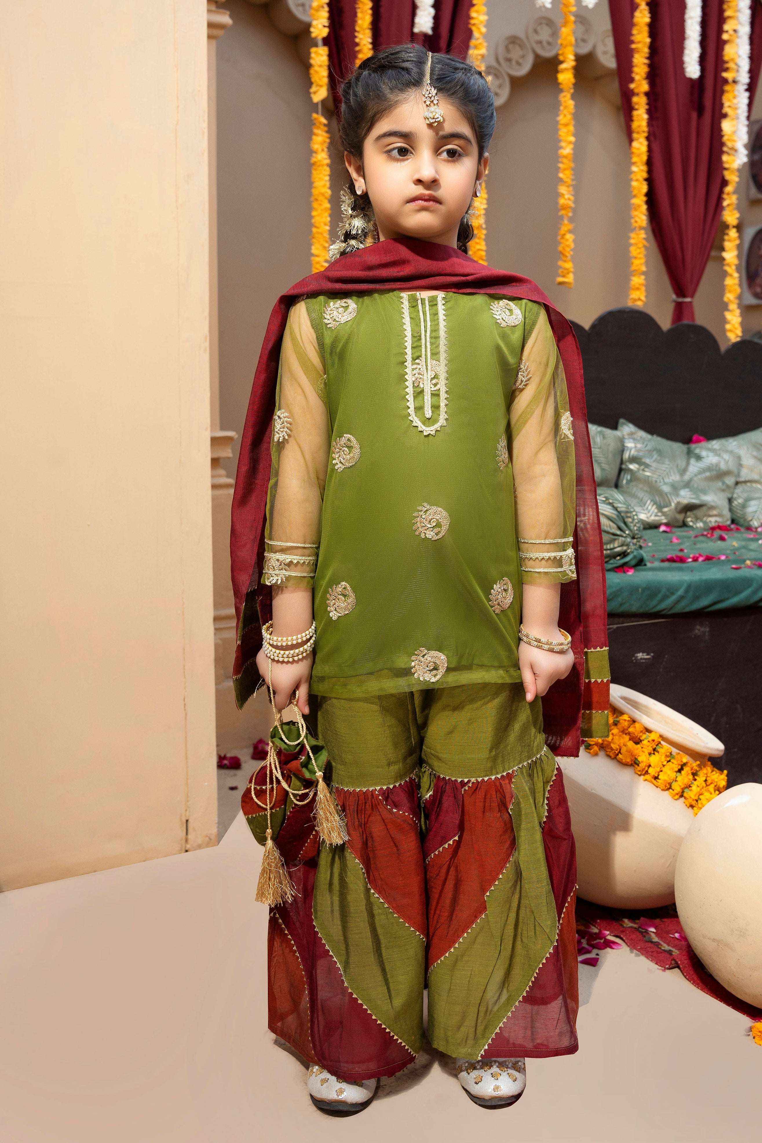 A young girl wearing a traditional 4-piece outfit with gold zari and sequins embroidery on a net shirt, chattapatti technique gharara in green, rust, and maroon with gold accents, and a matching dupatta with gold lace at the edges. She is also holding a potli.